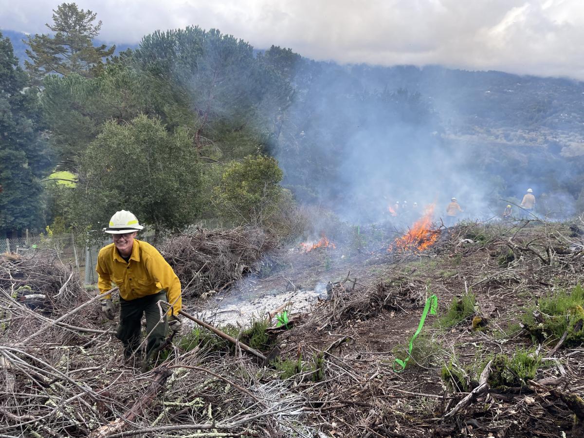  Student Zander Opperman is wearing a yellow fire resistant shirt and white hard hat while smiling in the foreground as be is preparing a pile to burn. In the midground several piles are in different stages of burning with complete burning and white ash, mid burning, and just ignited, with visible large flames. In the background other yellow shirt fire personnel are managing other piles, and in the distance the Santa Cruz Mountains are visible through the rising smoke.