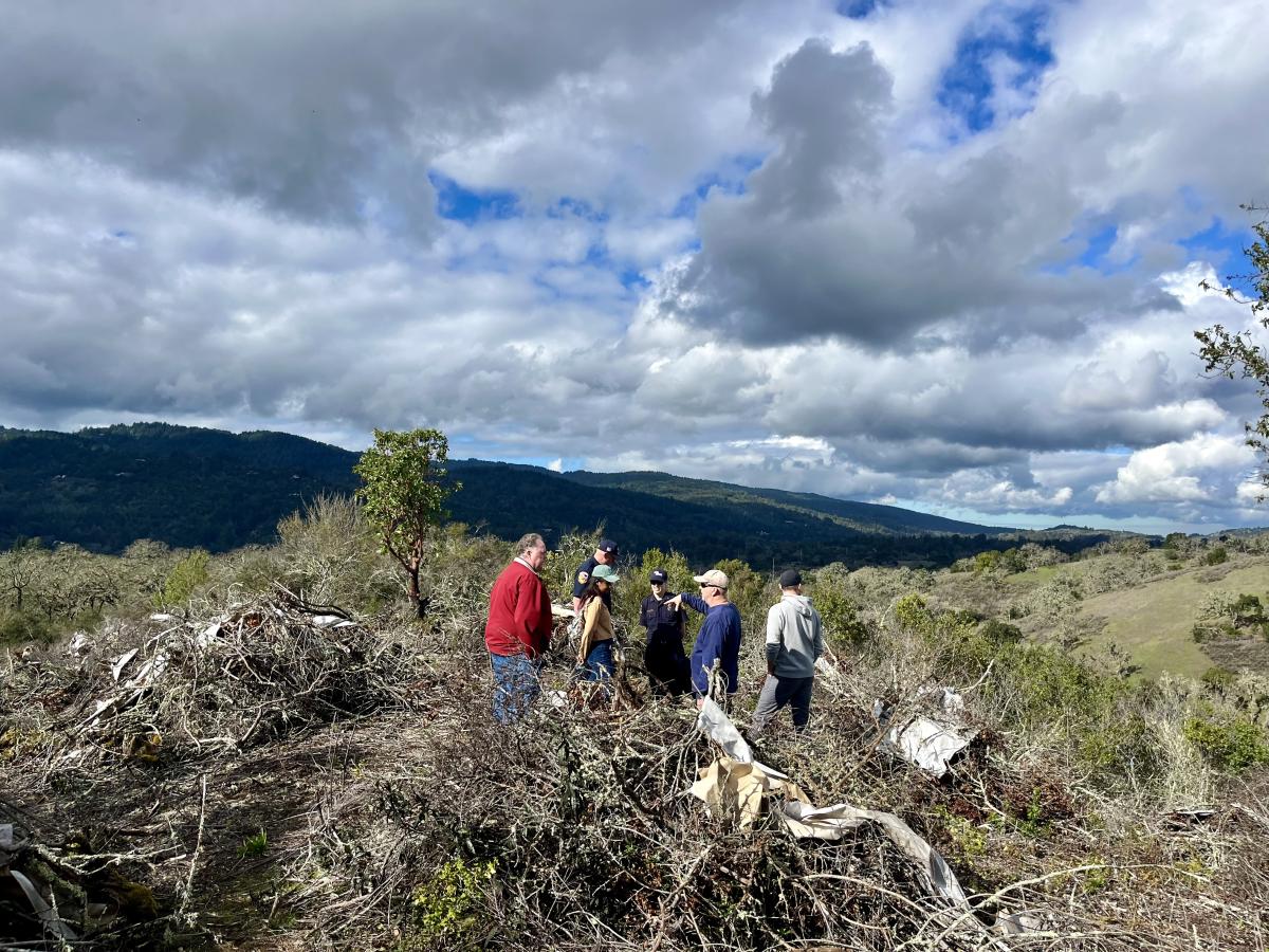 Group of 6 people standing and conversing among dried vegetation piles on top of a hill in a chaparral area. Santa Cruz Mountains are in distant background.