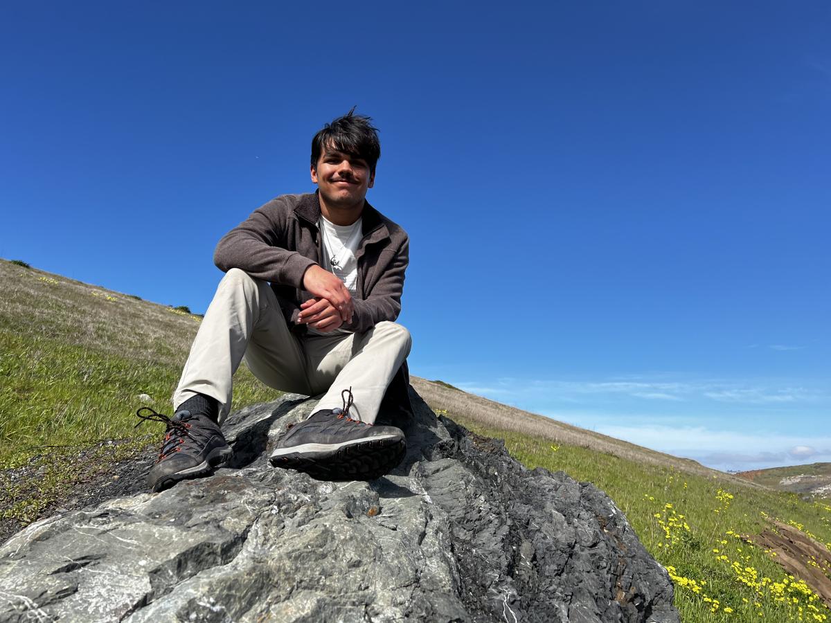 Nick Rodriguez sitting on a rock in the spring with flower field blooming behind him 